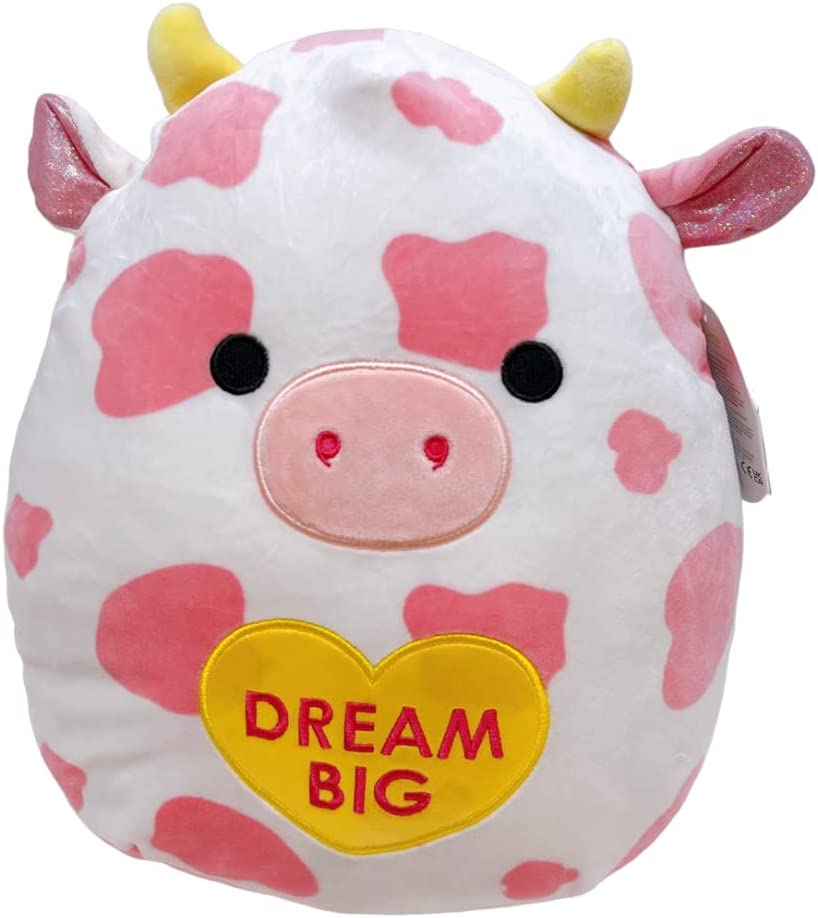 A pink and white cow Squishmallow holding a yellow love heart that says "dream big"