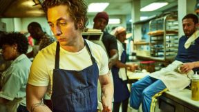 A chef in a blue apron looks despondent in a restaurant kitchen.