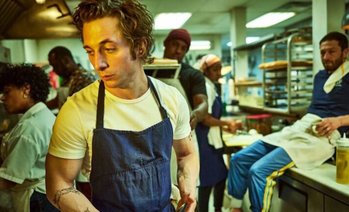 A chef in a blue apron looks despondent in a restaurant kitchen.