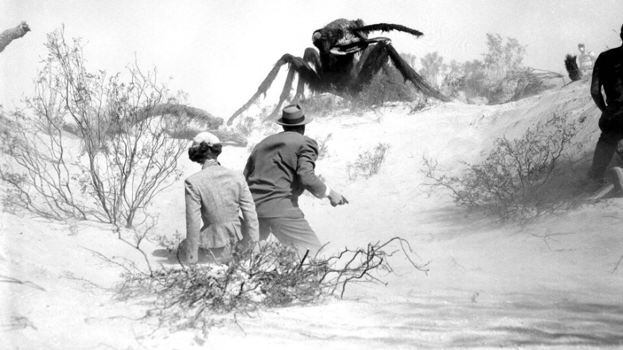 Black and white movie still from Them!, in which a man and woman look at a giant ant towering over them.