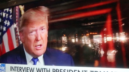 A Fox News graphic advertising an interview with Donald Trump is seen on a large TV, with bar tables seen in the screen's reflection.