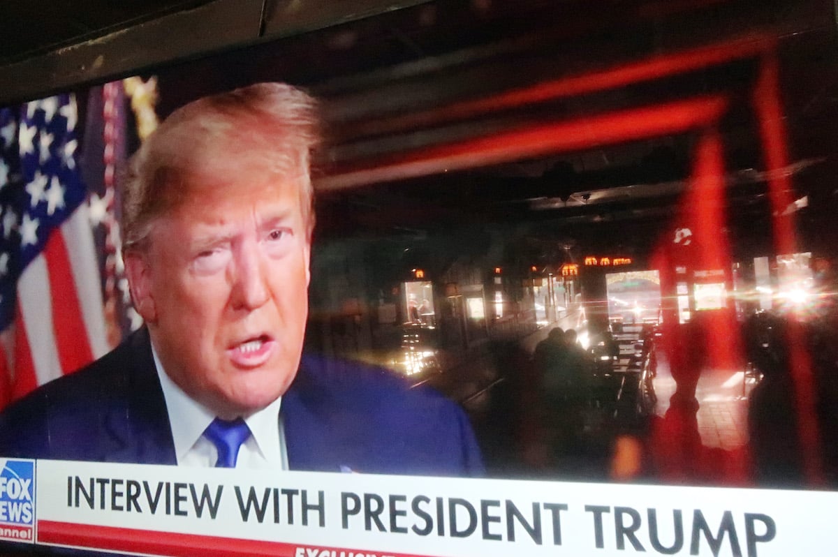 A Fox News graphic advertising an interview with Donald Trump is seen on a large TV, with bar tables seen in the screen's reflection.