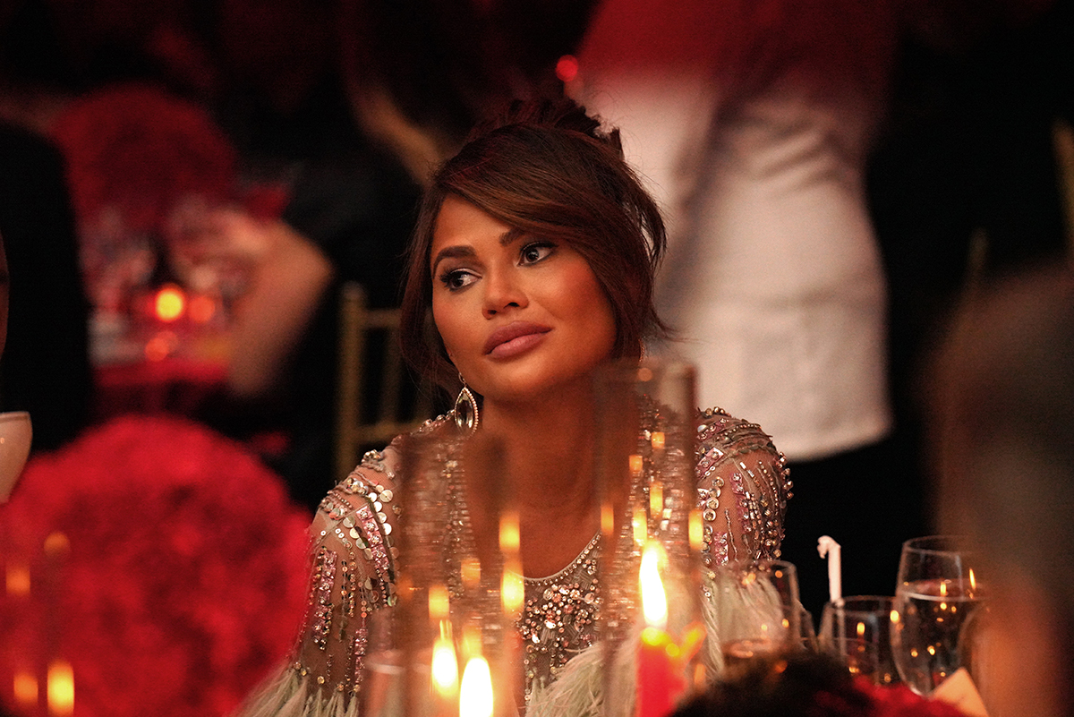 Chrissy Teigen stares into the distance sitting at a dinner table.