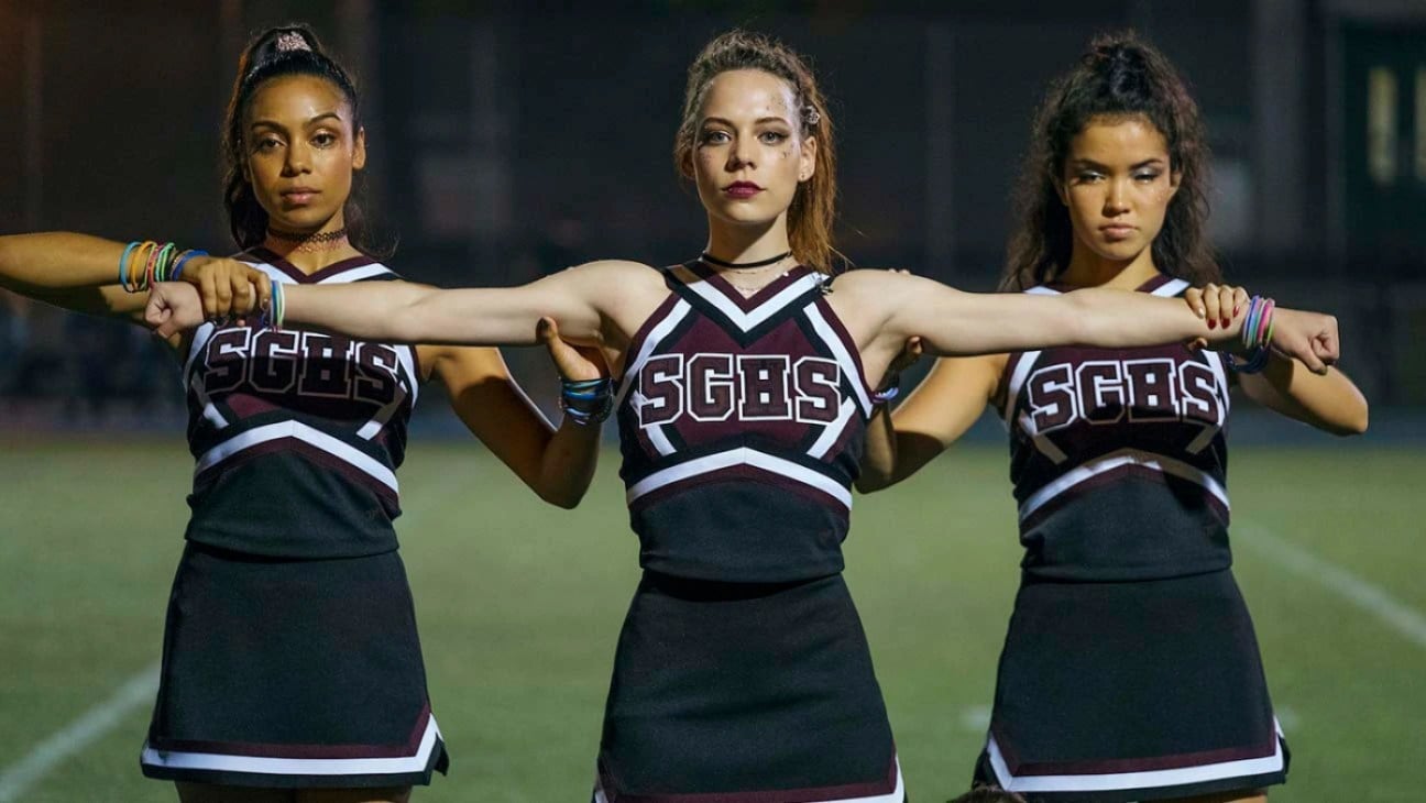 The cast of 'Dare Me' - three teen girls wearing cheerleading uniforms, standing on a football field