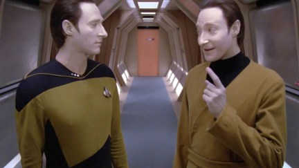 Two silvery white androids face each other mid conversation, one is confused and other is making a devious face