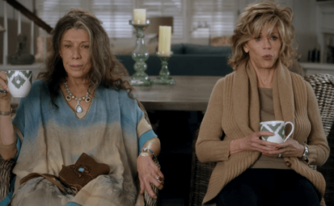 Grace and Frankie sit side by side on a porch holding mugs