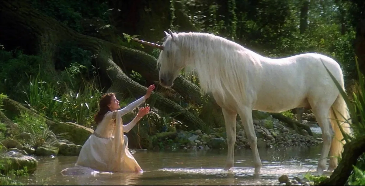 A woman kneeling down in front of a unicorn on the bank of a stream in 'Legend'