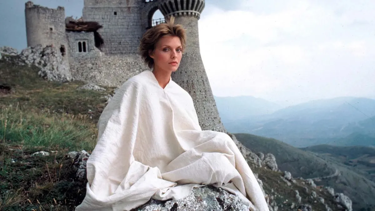 Michelle Pfeiffer as Isabeau in the film Ladyhawke, sitting on a rocky hillside in Central Italy, with a medieval castle in the background