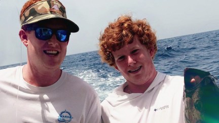 Two redheaded young adult/teen brothers smile with water behind them.
