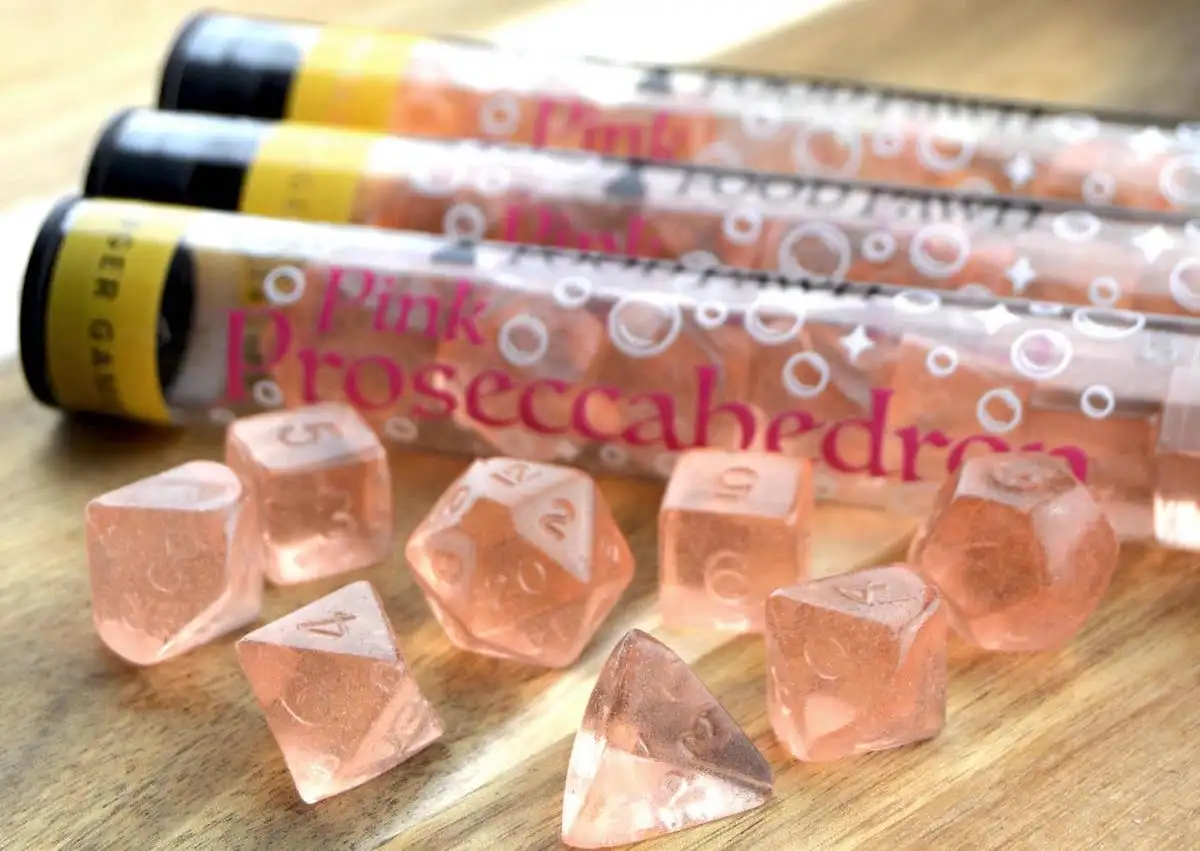 Test tube style packages full of pink dice sets with a handful scattered on the table in front of it
