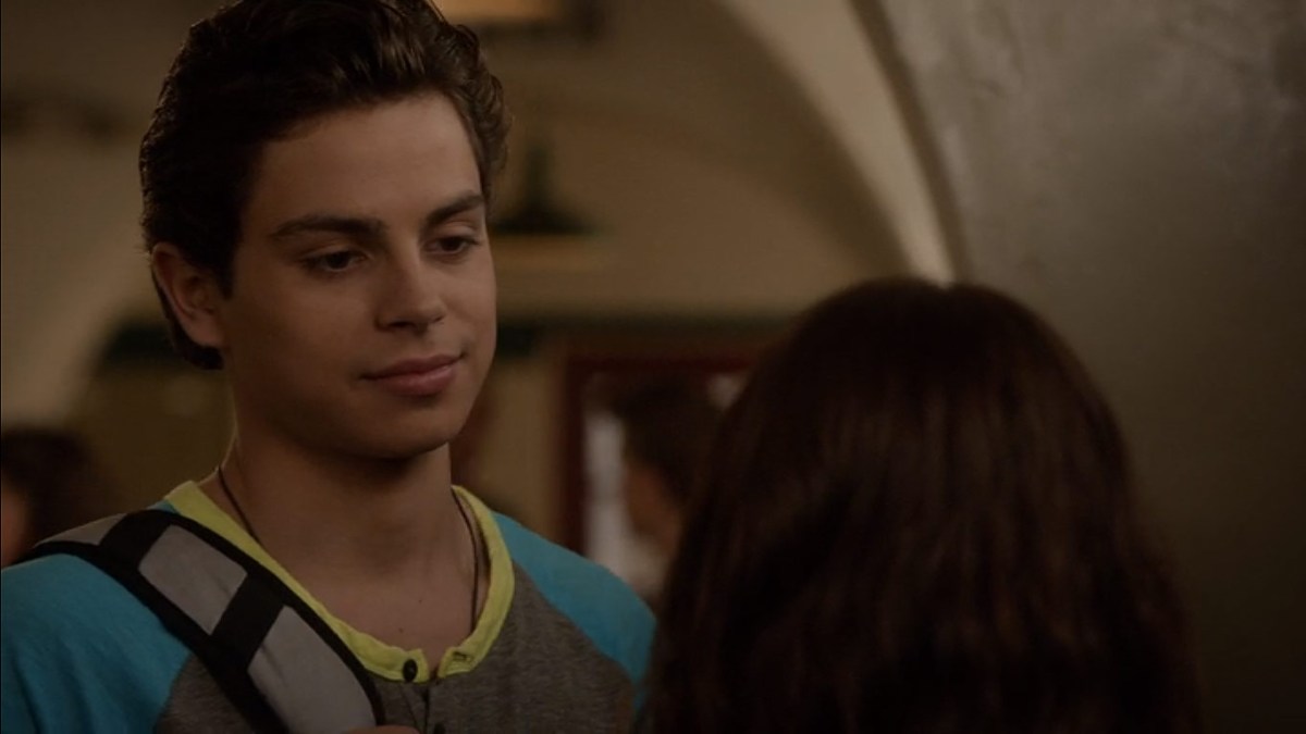 Jake T. Austin wears a dark t-shirt and a one strap backpack