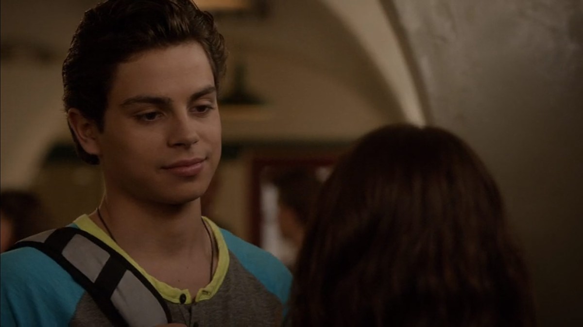 Jake T. Austin wears a dark t-shirt and a one strap backpack