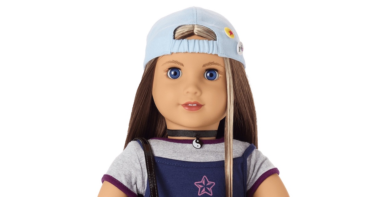 A close up of an American Girl doll with a backwards baseball cap, brown hair with a blonde streak by her face, and a yinyang necklace.