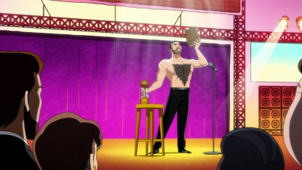 Brett Goldstein stands on a stage, shirtless. He holds up a book and has his hand on a statuette on a table.