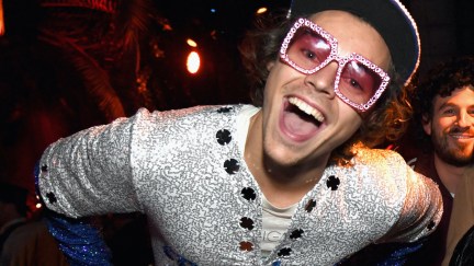 Harry Styles leans into the camera grinning, dressed is sparkly large sunglasses and a sparkly top.