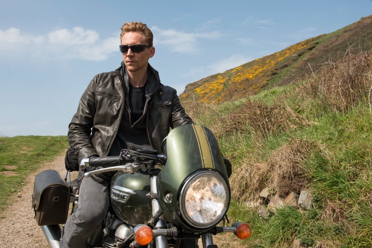 Tom Hiddleston as Jonathan Pine, sitting on a motorcycle wearing a leather jacket.