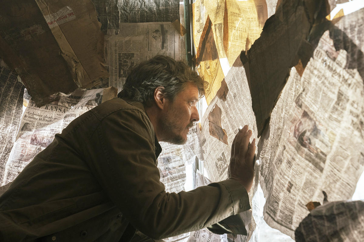 Pedro Pascal's Joel looks out of a window covered in newspapers in 'The Last of Us'
