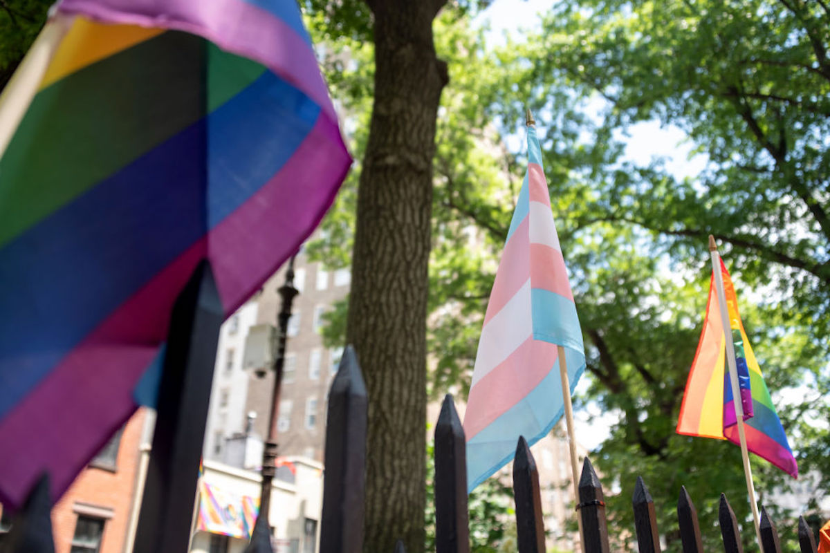 A Transgender Flag in between two Pride Flags outside with trees in the background.