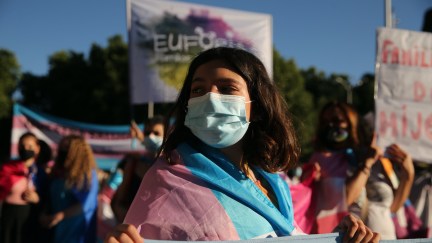 A young trans person wearing a face mask, draped in a trans flag, holds a sign at a protest.