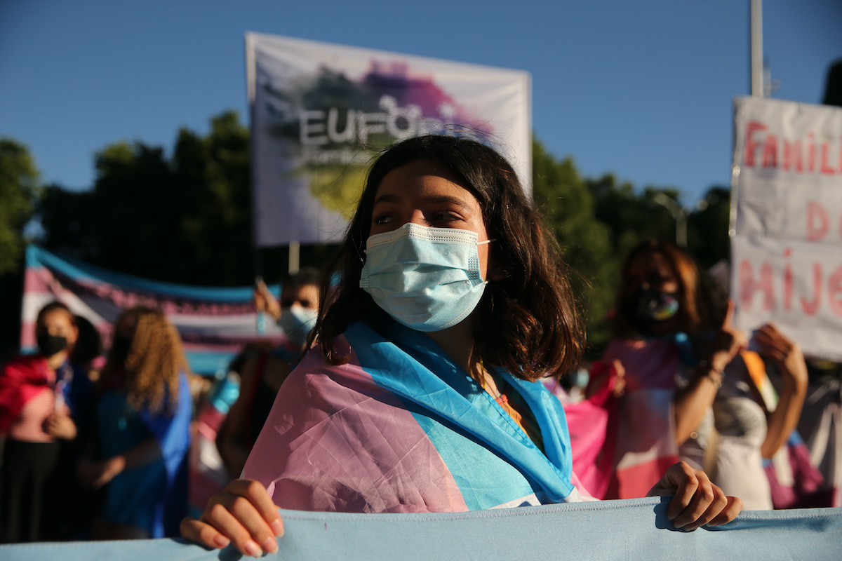 A young trans person wearing a face mask, draped in a trans flag, holds a sign at a protest.