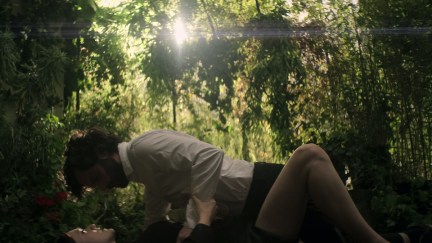 A man lies on top of a woman outdoors, surrounded by trees, as they look into each other's eyes.