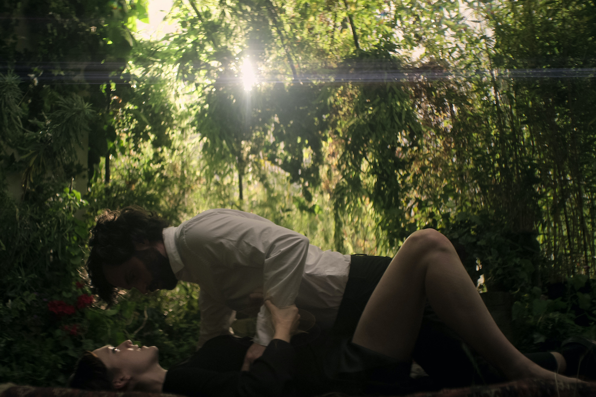 A man lies on top of a woman outdoors, surrounded by trees, as they look into each other's eyes.