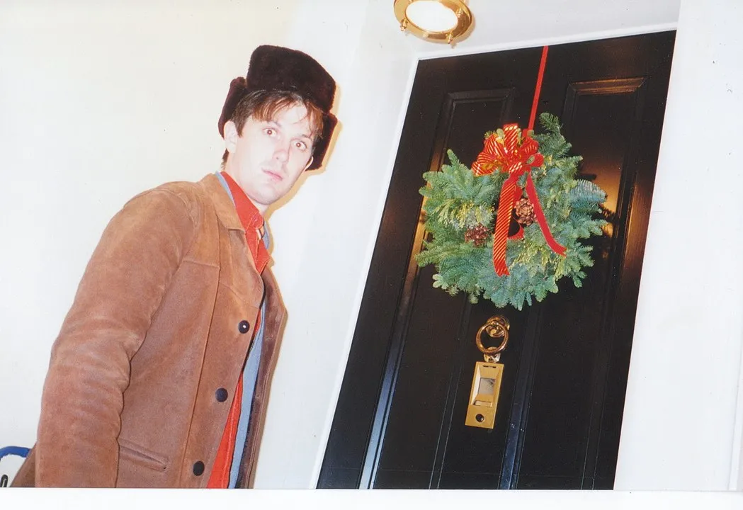 David McCormack in the 1990s, wearing a brown jacket and black hat and standing next to a door with a Christmas wreath on it.