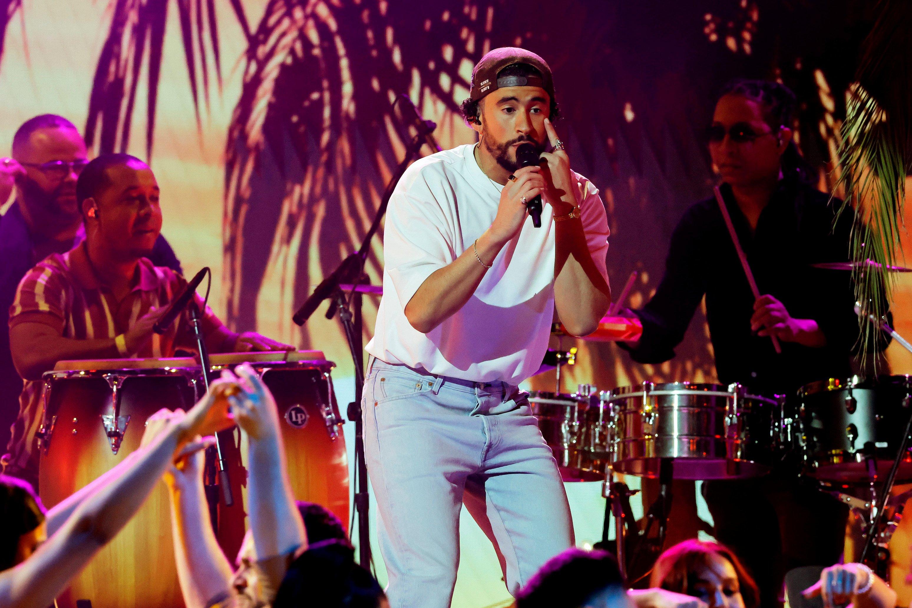 Image of Bad Bunny performing at the 23rd Annual Latin Grammys in Las Vegas. He is a light-skinned Puerto Rican man with a beard wearing a backwards, black baseball cap over his curly, black hair, a white t-shirt, and light blue jeans. He's singing into a microphone while pointing at his eye with the other hand. There are drummers behind him playing conga and snare drums, and there is a palm tree motif in the background under pink and orange light. 