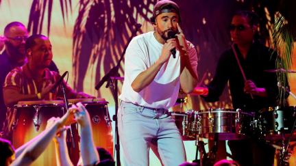 Image of Bad Bunny performing at the 23rd Annual Latin Grammys in Las Vegas. He is a light-skinned Puerto Rican man with a beard wearing a backwards, black baseball cap over his curly, black hair, a white t-shirt, and light blue jeans. He's singing into a microphone while pointing at his eye with the other hand. There are drummers behind him playing conga and snare drums, and there is a palm tree motif in the background under pink and orange light.