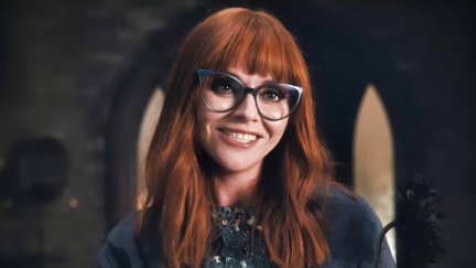 Christina Ricci as Marilyn Thornhill on Netflix's 'Wednesday.' We see her from the shoulders up, and she's smiling a big smile. She has long, red hair with bangs, wears blue, cat eye glasses and a greyish top.