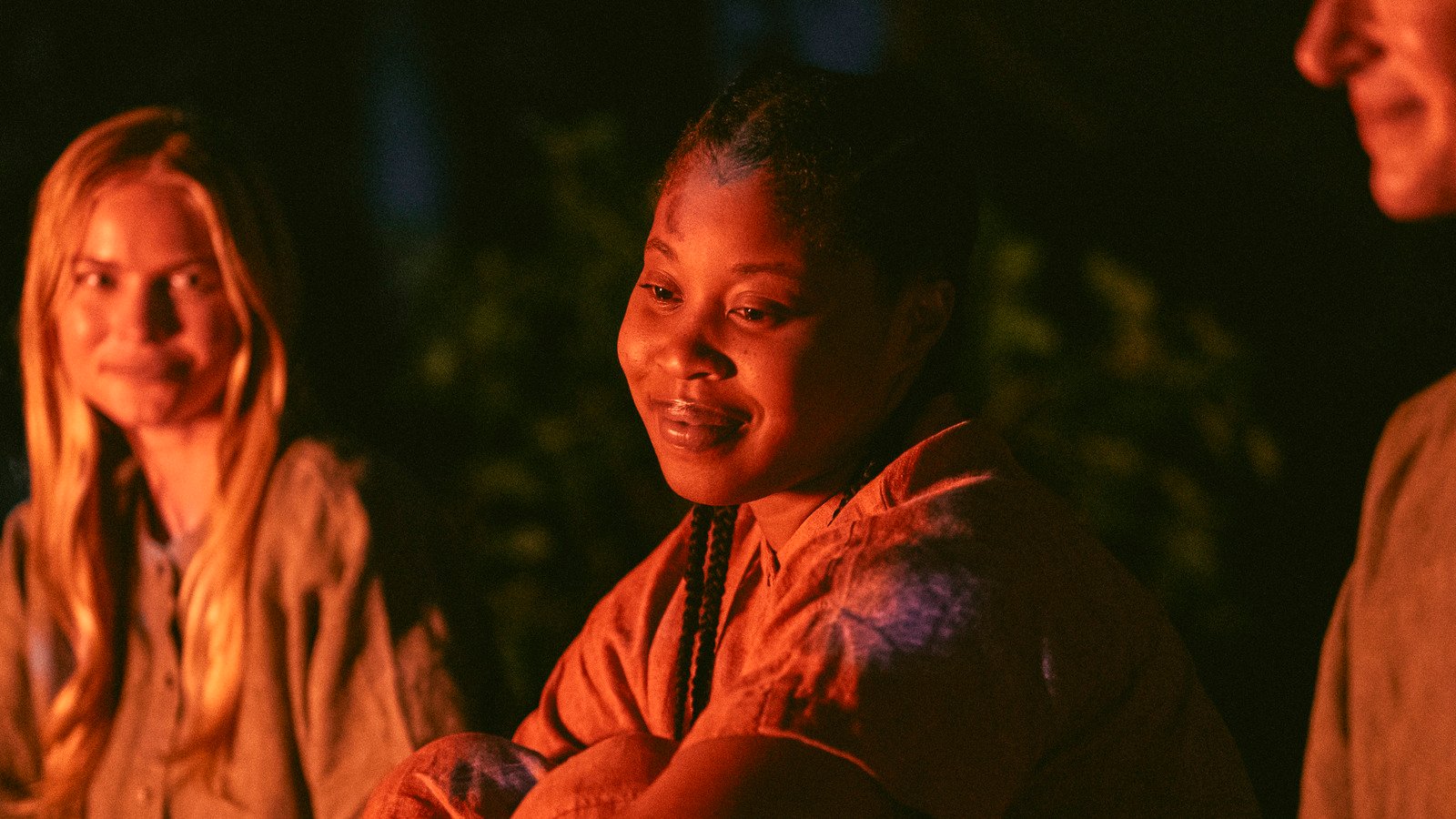 Image of Dominique Fishback as Dre in Amazon's 'Swarm.' We see Dre from the shoulders up as she's seated around a fire with two other women who are slightly out of focus. She is lit by firelight, and has a soft smile on her face as she looks at the fire. She's hugging her knees. She has long, dark braids.