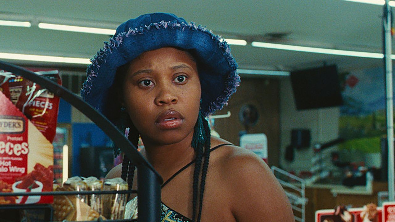 Image of Dominique Fishback as Dre in Amazon's 'Swarm.' She is standing in a grocery store, visible from the shoulders up as she stands next to a shelf holding snacks. her shoulders are bare, and she has long, black braids coming out from under a wide-brimmed, blue hat. She's looking into the distance.
