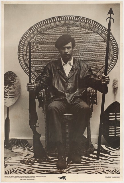 A poster of Huey Newton sitting in a rattan throne chair wearing a beret and a black leather jacket while holding a shotgun in his right hand and a spear in his left hand. 