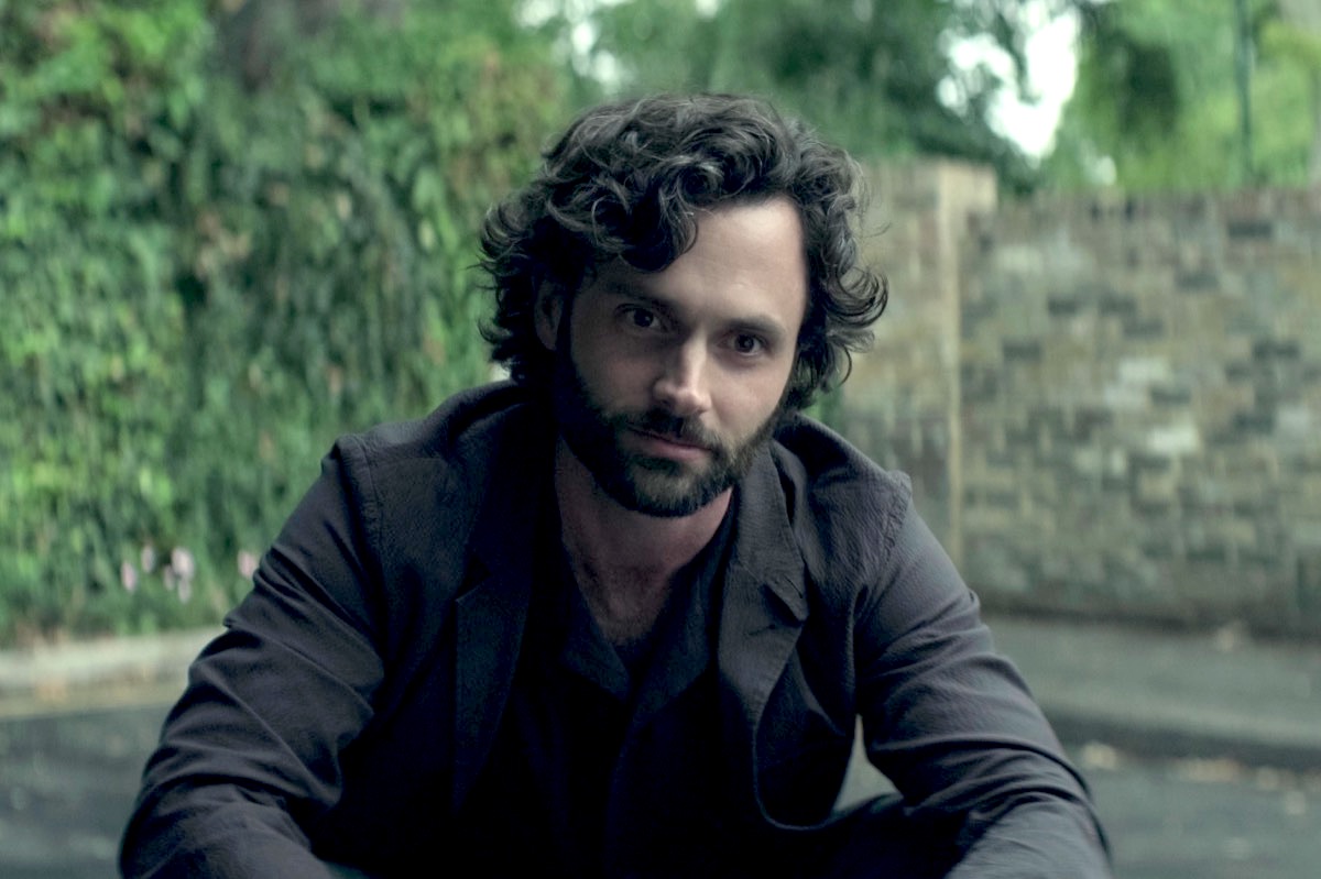 A man with short curly hair kneels in a park and stares into the camera with a deceiving look.