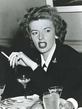 Marjorie Cameron smokes a cigarette at a table. The picture is clearly from the mid-20th century. Cameron is wearing a black jacket.