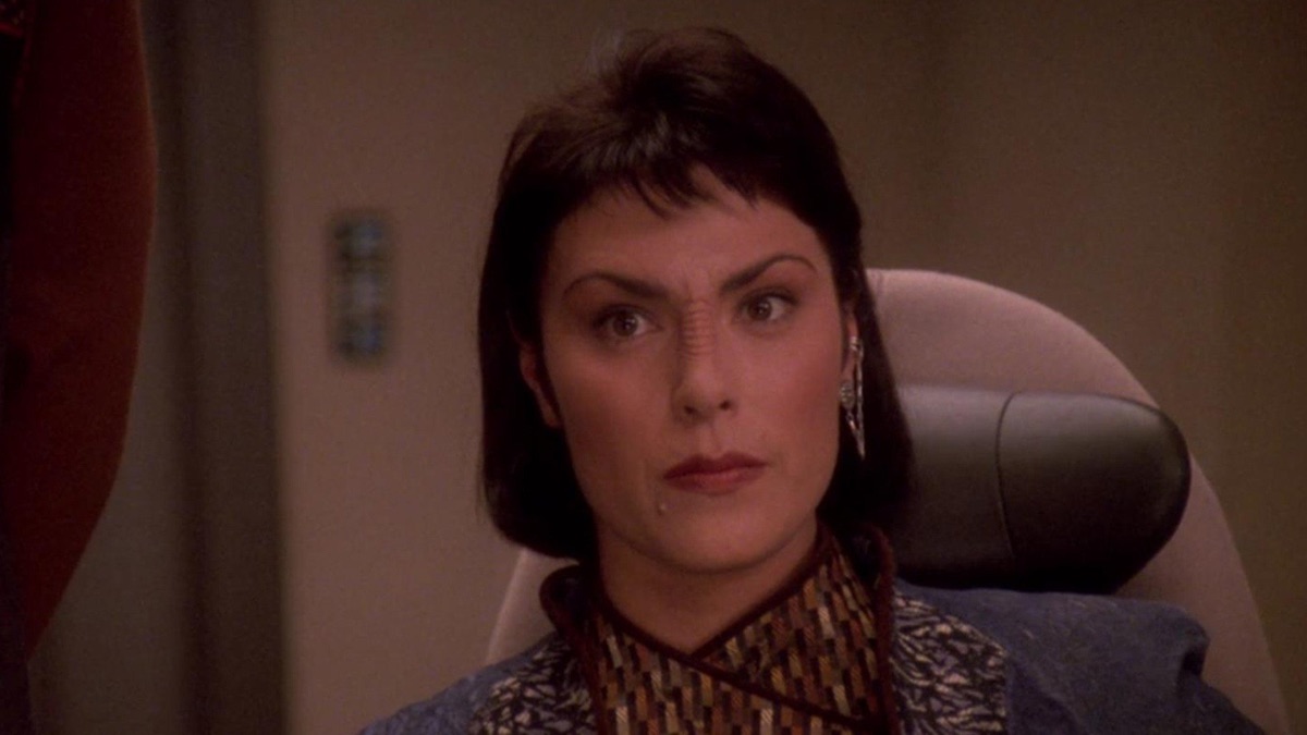 Image of Michelle Forbes as Ensign Ro Laren on "Star Trek: The Next Generation." She's seated in a chair on the Enterprise and visible from the neck up. She's dressed as a Maquis, with a high, purple and gold collar visible at her neck. Her dark, chin-length hair has bangs. Her Bajoran cuff earring is visible on her left ear (our right). 