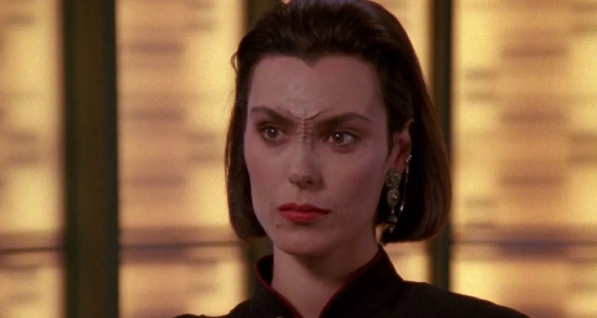 Image of Michelle Forbes as Ensign Ro Laren on "Star Trek: The Next Generation." We see her from the neck up. She's standing on a transporter pad. She has chin-length, dark hair that's brushed back and she has a widow's peak. She has Bajoran nose-ridges on the bridge of her nose, and a Bajoran cuff earring on her left ear (our right). She wears red lipstick and has a mole on the right side of her mouth. The dark collar of a Starfleet uniform is visible at her neck.