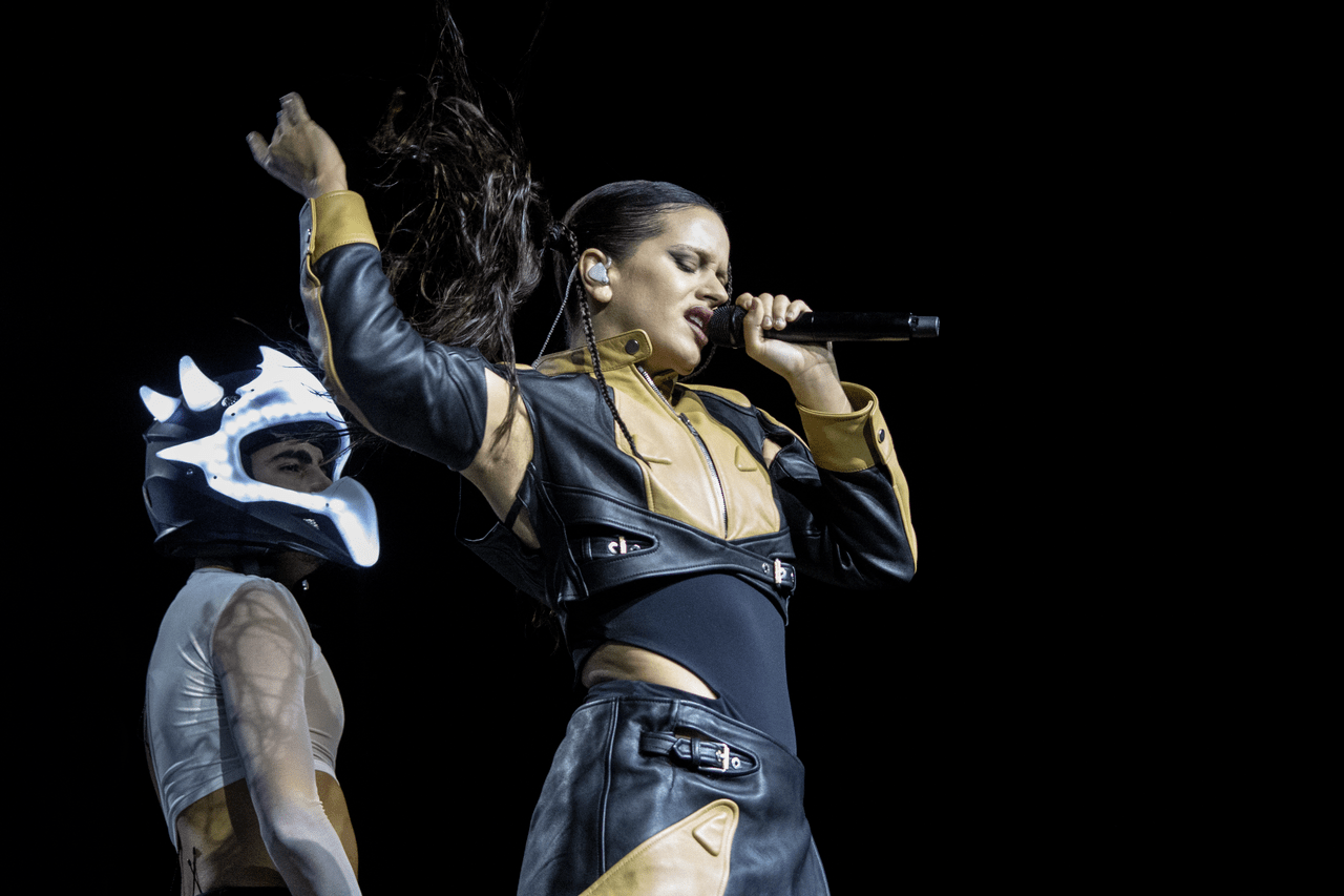 Image of Rosalía singing during a concert in Chile. She is a white, Spanish woman dressed in a black and yellow leather jacket and pants with a visible black leotard underneath that shows skin on her waist and under her arm. She's holding a microphone in one hand and flipping her long, dark hair with the other. She's singing with her eyes closed and there's a monitor in her ear. A person in a motorcycle helmet with horns on it stands behind her. 