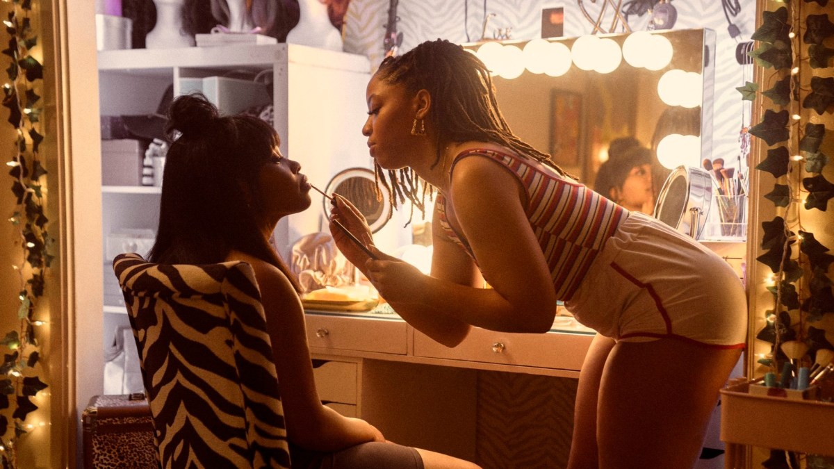 Image of Dominique Fishback as Dre and Chloe Bailey as Marissa in Amazon's 'Swarm.' They are in front of a vanity table with large lights around the mirror. Dre is seated in a zebra-print chair with her eyes closed as Marissa does her make-up. Her hair is black and shoulder-length. She has bangs, and some of her hair is pulled up into a bun at the top of her head. Marissa is bent over as she applies Dre's lipstick. She has long braids that go down to the middle of her back, and she's wearing gold doorknocker earrings. She's wearing a red, blue, and white striped tank top and white short shorts with red piping.