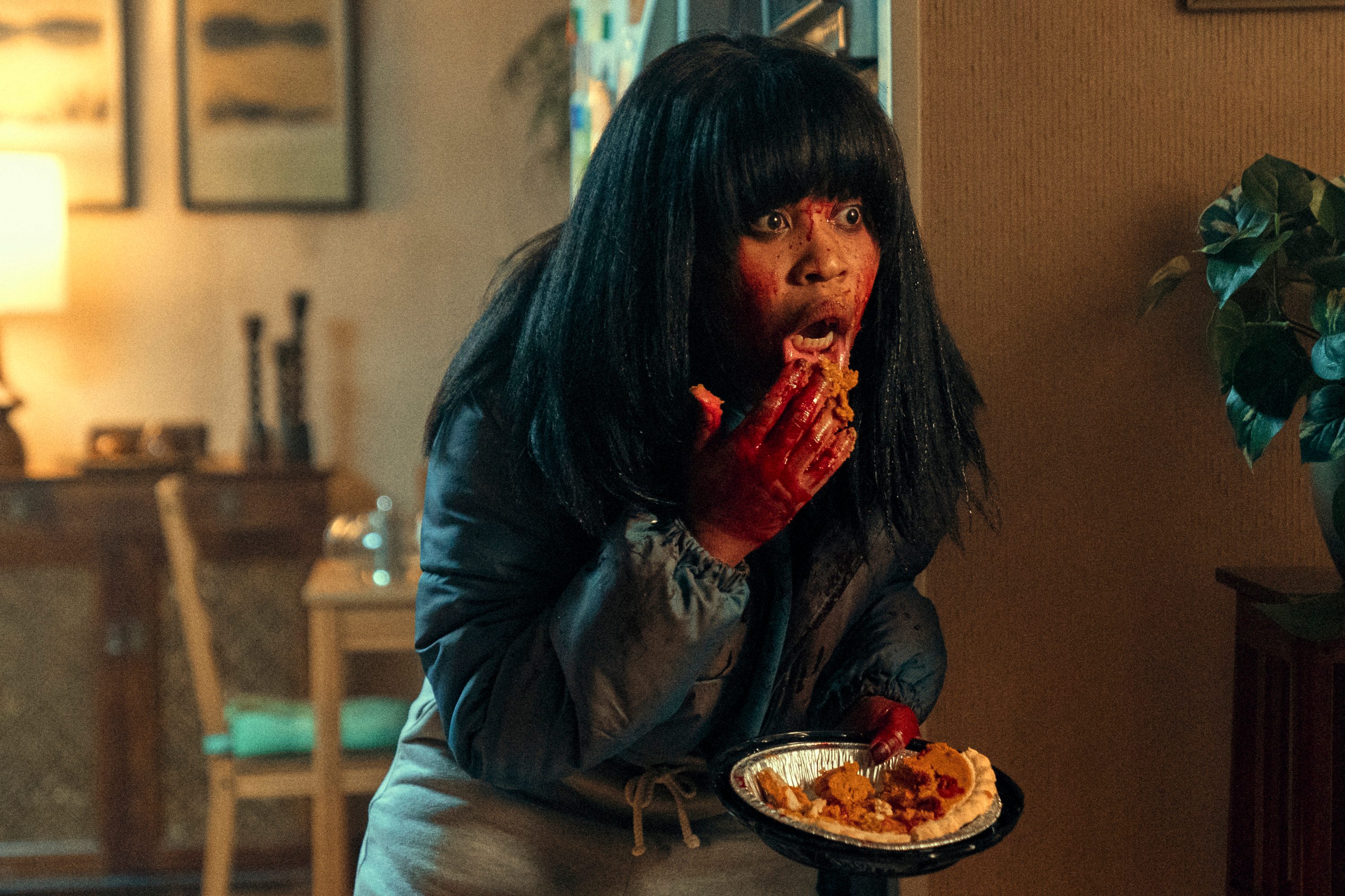 Image of Dominique Fishback as Dre in Amazon's 'Swarm.' She is a Black woman, and is standing in a house with her face and hands covered in blood as she holds a half-eaten pumpkin pie and scoops some into her mouth with her hand. She has long, black hair and bangs, and wears a puffy, black jacket and grey sweatpants.