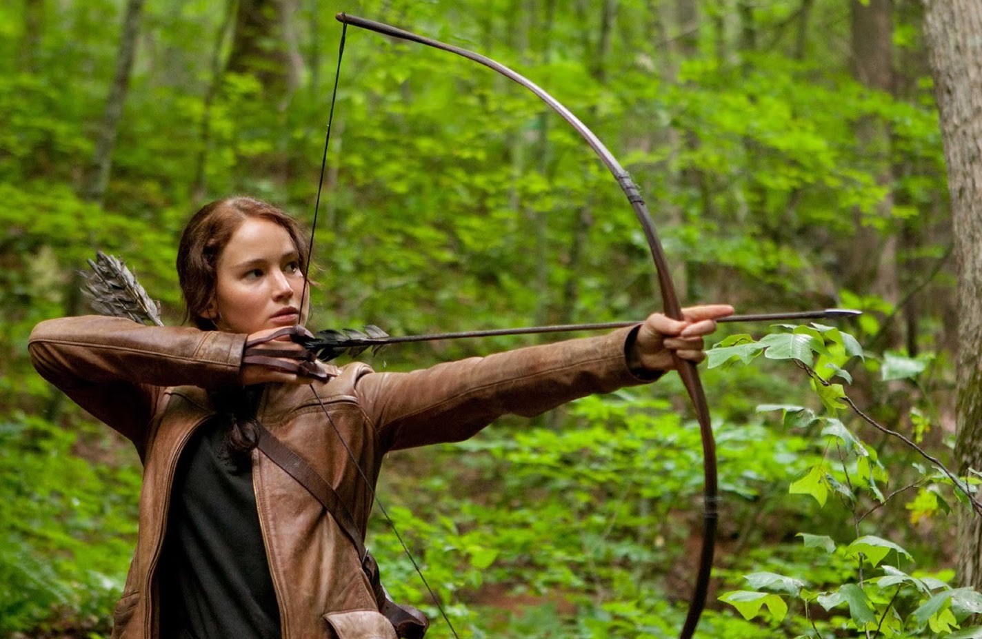 A brown haired teen girl with her hair in a braid pulls back her bow and arrow in the forest.
