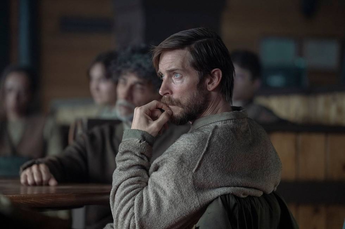 Image of Troy Baker as James in HBO's 'The Last of Us.' James is inside a restaurant seated at a table with other people. He has short, brownish-reddish hair, and a beard and wears a greenish inside-out sweatshirt with a green jacket draped on the chair behind him. His elbow is on the armrest, and that hand is fidgeting with his beard as he listens to someone off to the left. 