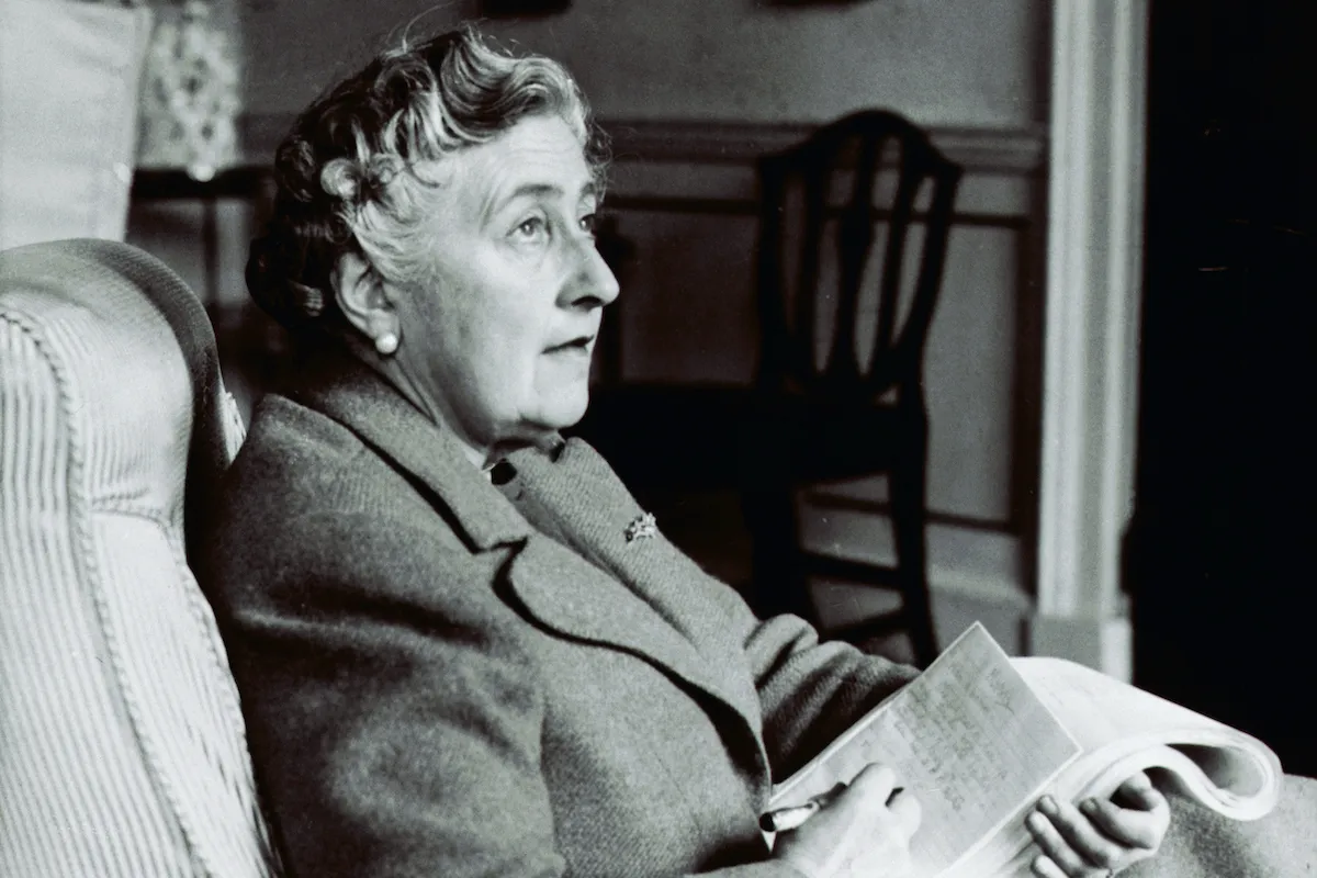 A black and white photo of Agatha Christie sitting, holding a stack of papers, staring off wistfully