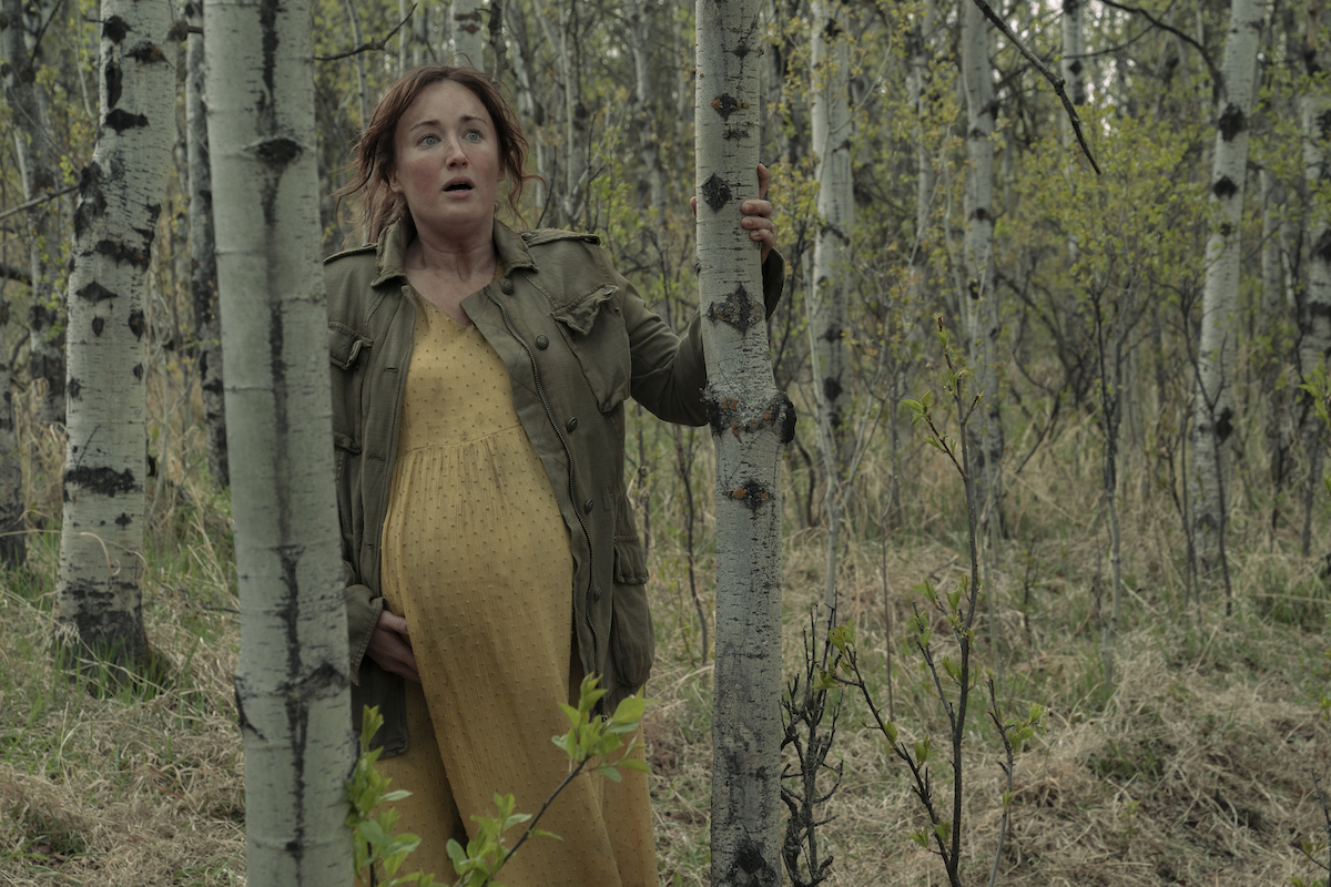 A pregnant woman in a yellow dress and leather jacket leans against a tree in the woods, looking frightened.