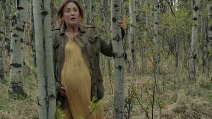 A pregnant woman in a yellow dress and leather jacket leans against a tree in the woods, looking frightened.