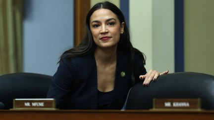 Alexandria Ocasio-Cortez smiles standing behind a desk in the House.