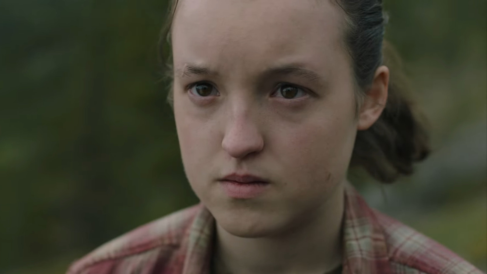 Image of Bella Ramsey as Ellie on 'The last of Us' on HBO. Close-up image of Ellie, who is outdoors and looking at something with a serious expression. Her dark hair is pulled back into a pony tail, and she's wearing a red plaid collared shirt. 