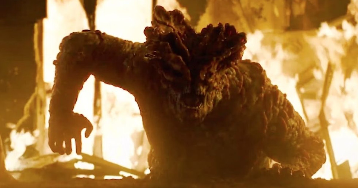 Close-up image of a bloater infected from HBO's the last of us. It is emerging from the ground and everything around it is on fire. It is enormous and covered in fungal plates. It's mouth is open.