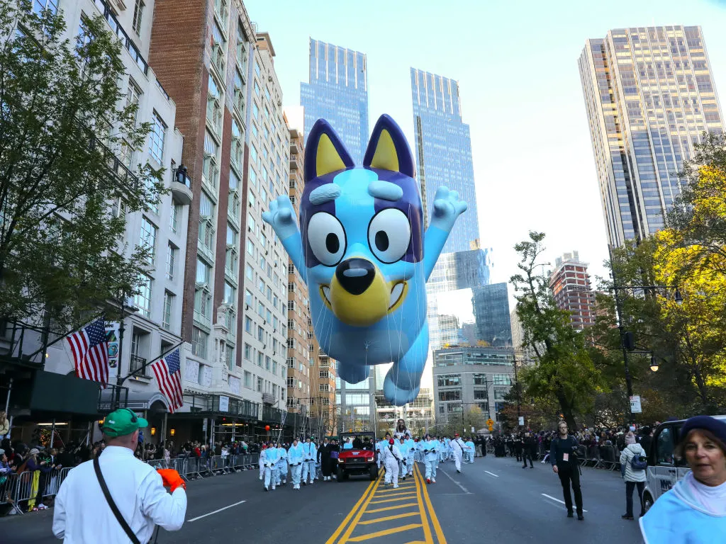 A gigantic Bluey balloon floats down the street, pulled by people wearing white, in the Macy's Thanksgiving Day Parade. 