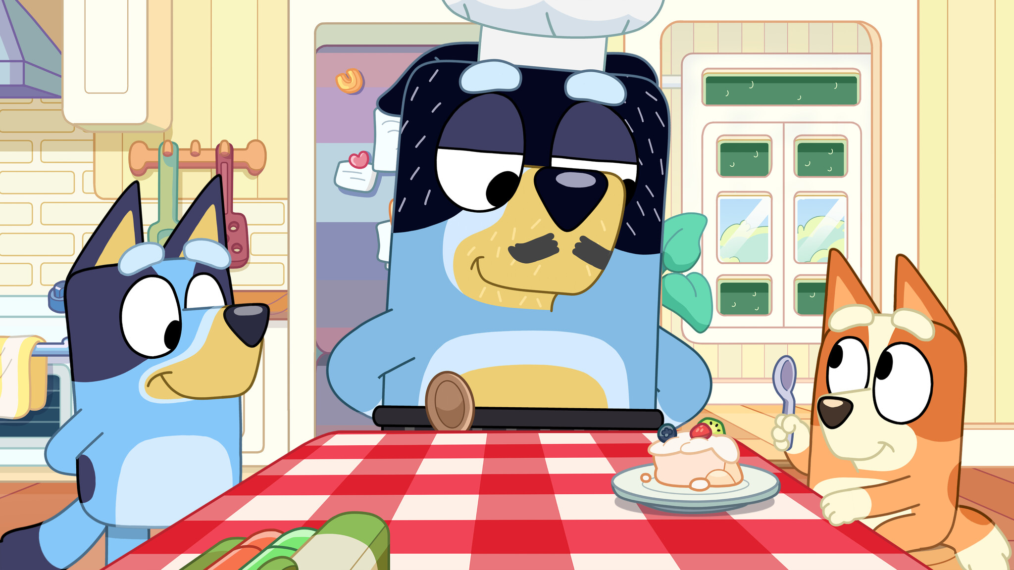 Bandit wears a chef's hat and fake mustache while Bluey and Bingo sit at the kitchen table. Bandit smiles down at Bingo.